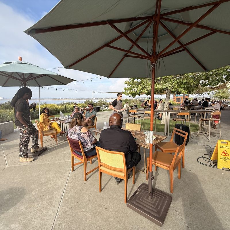 Neighbors talking around tables under large umbrellas at an outdoor community gathering.
